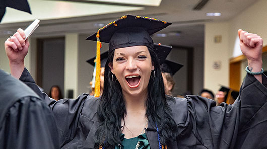 An NMC graduate in cap and gown raising her hands and smiling during NMC's 2026 Commencement