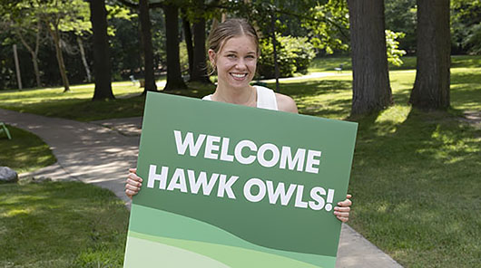 A smiling NMC residential advising student holds a Welcome Hawk Owls sign