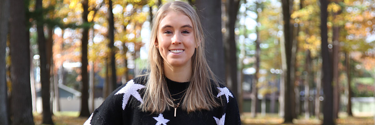 An NMC student in a sweater standing under the pines on NMC's Front Street campus