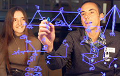 An NMC Engineering Program instructor demonstrates a calculation on a transparent whiteboard while an engineering student looks on